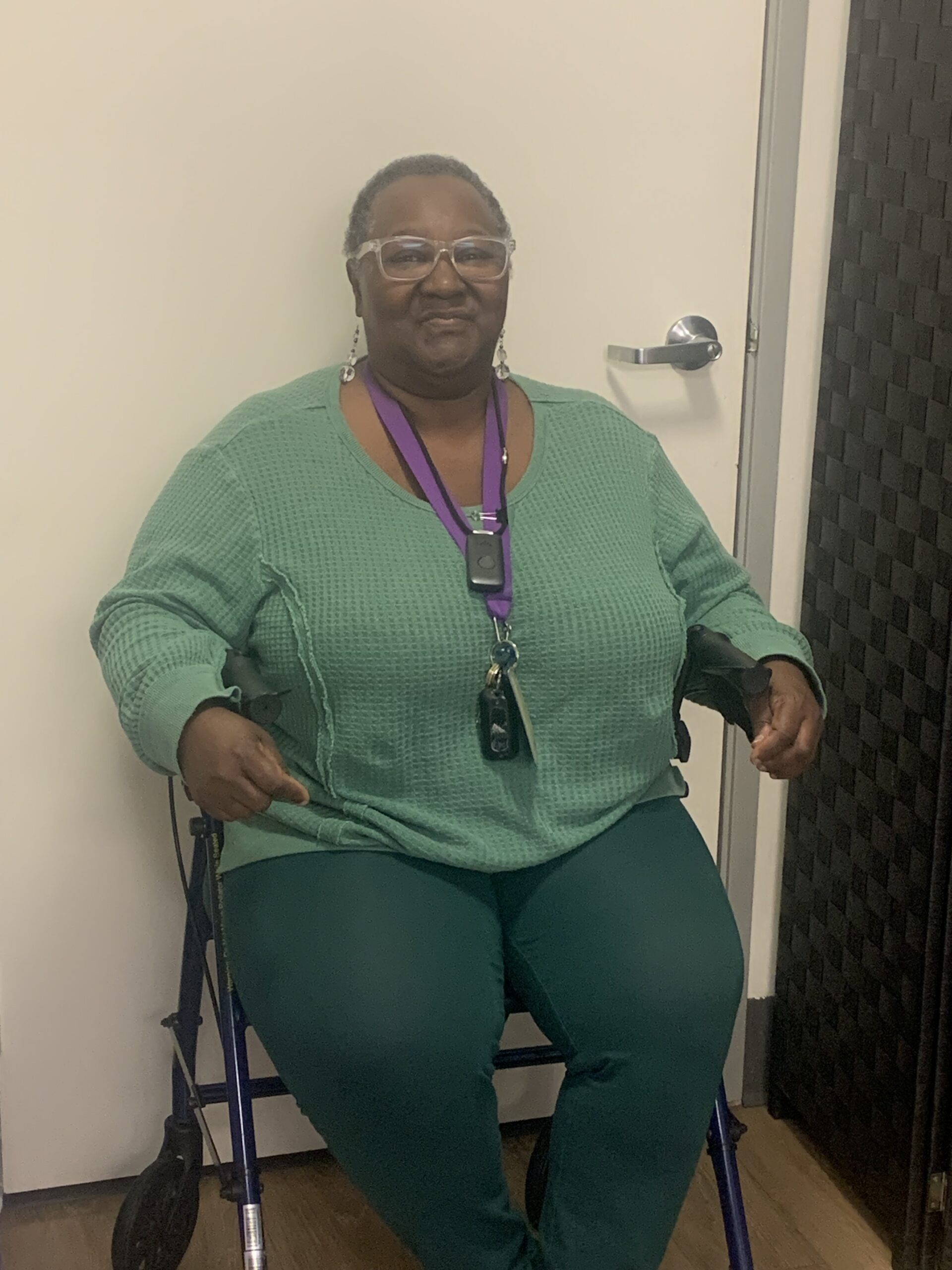 Cynthia wearing a long sleeve green top and dark green pants. She has short, dark hair and glasses. Smiling while sitting in her walker.