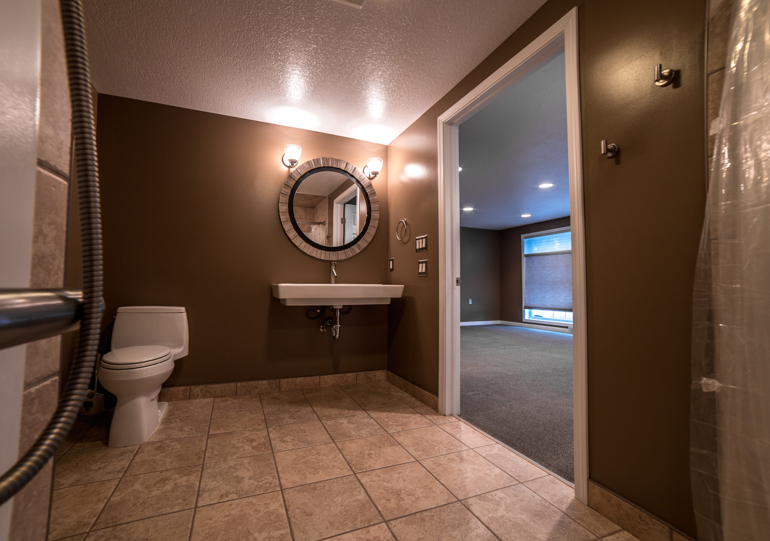 Picture of open bathroom. Brown painted walls, with circular mirror above stink with open space underneath. Wide door on the right, with toilet to the left.