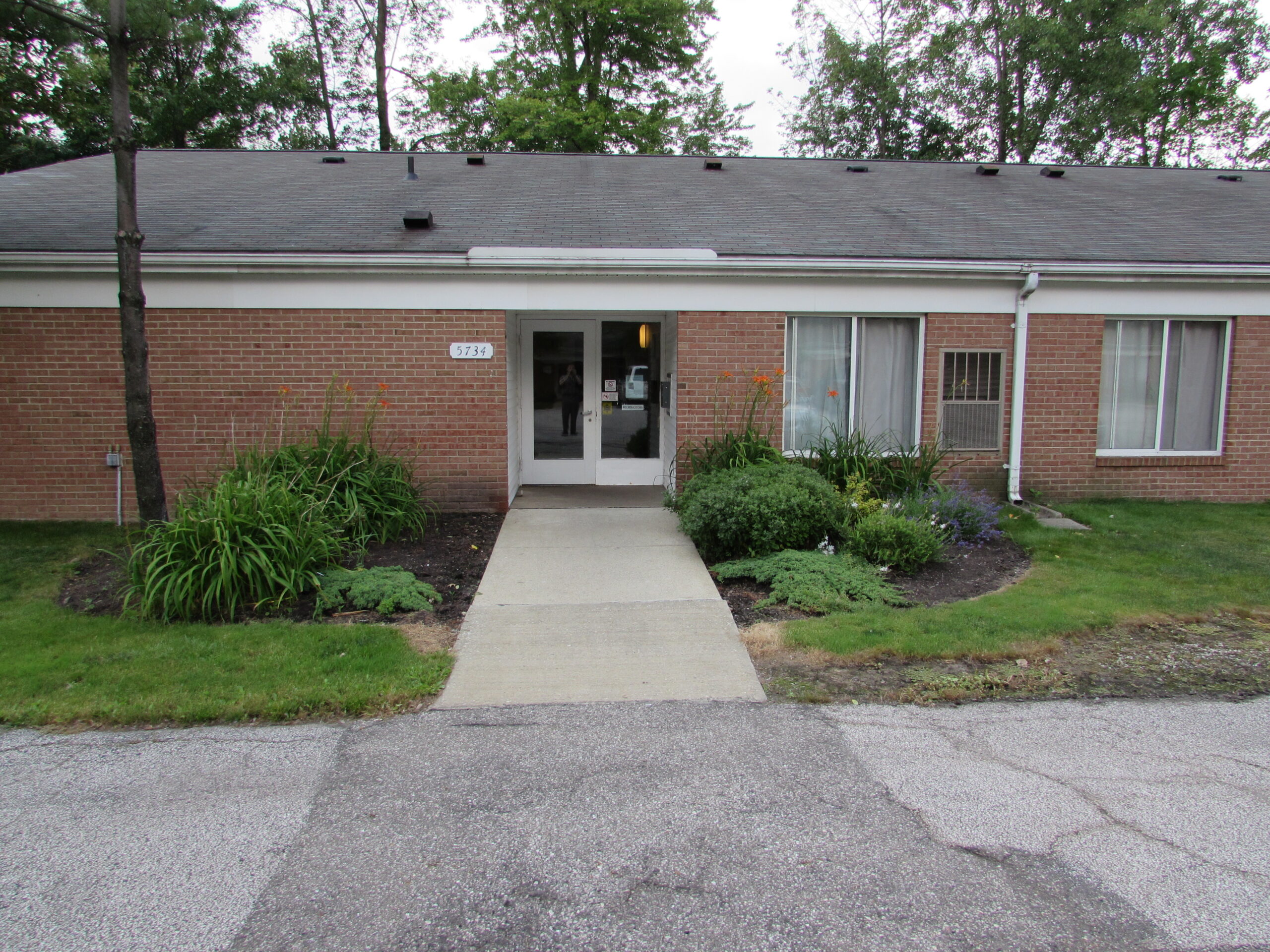 Wide sidewalk up to white doors. Greenery on both sides of door.
