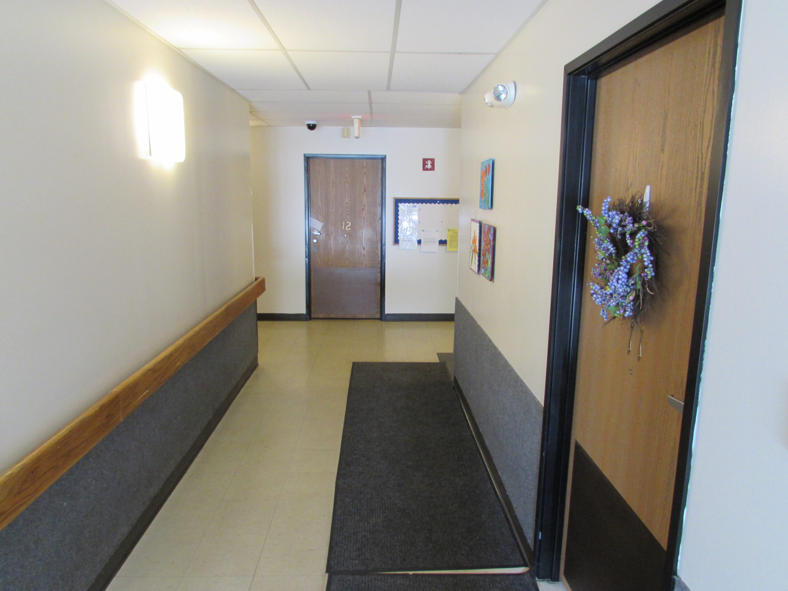 Wide hallways with grab bar on the left of the hallway. Apartment door with wreath on the right and apartment door at the end of the hallway.