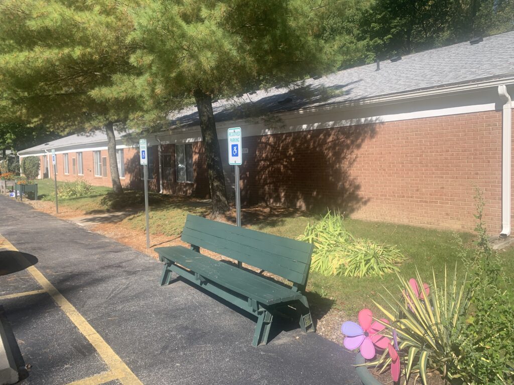 [Image] Close up picture of green bench in front of Lake Vistas building. Bench in front of Handicap parking sign, next to flower pot.