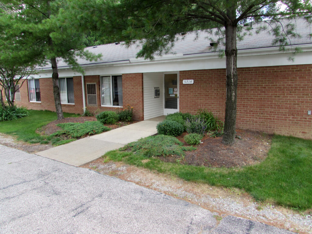 Image of Lake Vistas. One story building with brick on the outside and white siding. Walkway up to the front door with bushes outside the building.