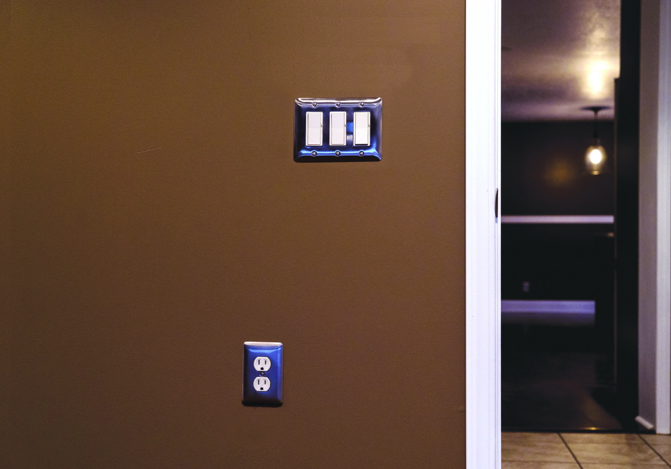 Picture of painted brown wall with hallway to the right. Three paddle light switches and lowered outlet.