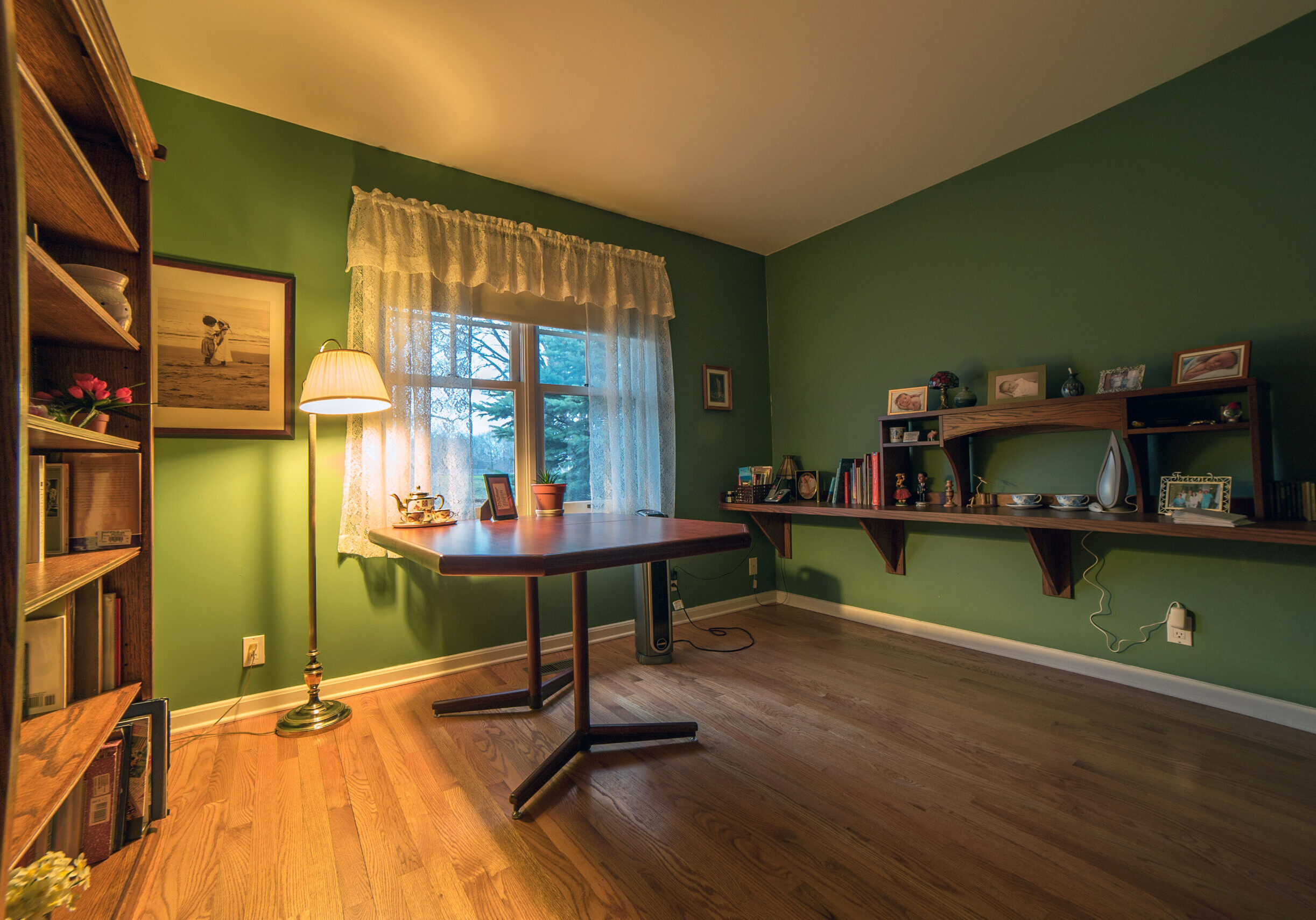 Picture of office in home. Green walls with white curtains, tall bookshelf on the left, wood shelving on the right with books and pictures. Table in the middle of the room, task lighting throughout and open space to maneuver.